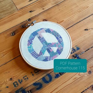 May include: A white embroidery hoop with a colourful peace sign design. The peace sign is made up of different coloured squares and rectangles. The hoop is on a wooden surface. PDF Pattern Cornerhouse 115