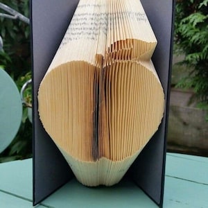 May include: A book folded into the shape of a heart, standing upright on a light blue wooden surface. The book is open and the pages are folded to create the heart shape. The book is bound in black fabric.