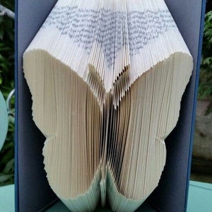 May include: A book folded into the shape of a butterfly. The book is open and the pages are folded to create the wings of the butterfly. The book is sitting on a light blue surface.