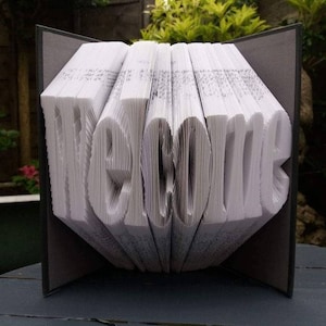 May include: A book art sculpture that has been folded to create the word "welcome". The book is open and the pages are folded to create the letters. The book is sitting on a wooden table.