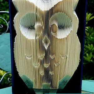 May include: A book folded into the shape of an owl, with a blue cover and a white background. The book is standing on a teal table. The text "PDF Pattern Cornerhouse 115" is visible in the bottom right corner of the image.