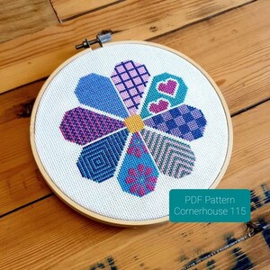 May include: A colorful cross-stitch pattern of a flower with a patchwork design. The pattern is in a wooden embroidery hoop and includes the text "PDF Pattern Cornerhouse 115".
