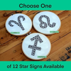 May include: Three embroidery hoops with black and white star sign designs. The hoops are on a wooden surface. The signs are Aries, Leo, and Sagittarius.