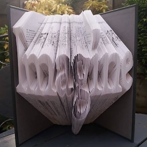 Puede incluir: Una escultura de arte de libro, doblada en la palabra "remarkable", se muestra sobre una superficie gris. El libro es blanco con texto negro y está encuadernado en una cubierta gris.