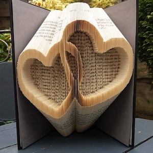 Op de afbeelding: Een boekkunst sculptuur met twee harten gevouwen uit de pagina's van een boek. Het boek is gebonden in grijze omslagen en staat op een grijs houten oppervlak.