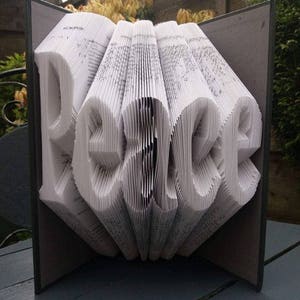 May include: A book art sculpture of the word "peace" created by folding the pages of a book. The sculpture is displayed on a gray wooden table.
