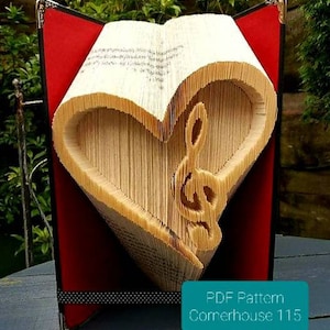 May include: A handcrafted book sculpture with a heart-shaped void and a treble clef. The pages are folded to create the design, set against a red background. The text "PDF Pattern Cornerhouse 115" is visible. The piece is a creative decorative item.