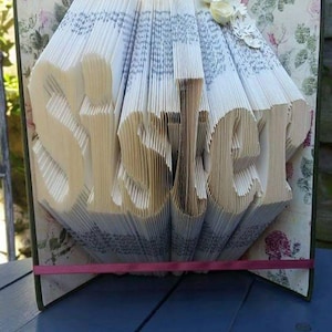 Peut inclure: Une sculpture d'art de livre avec le mot "Sœur" plié dans les pages. Le livre est recouvert d'un motif floral et est orné d'un ruban rose.