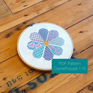 May include: A cross-stitch pattern of a flower with a yellow centre and petals in shades of blue, purple, and pink. The pattern is in a round embroidery hoop. The text "PDF Pattern Cornerhouse 115" is below the hoop.