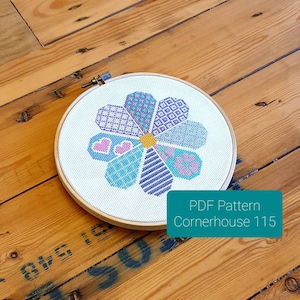 Op de afbeelding: Een kruissteekpatroon van een bloem met een geel centrum en bloemblaadjes in tinten blauw, paars en roze. Het patroon is in een ronde borduurring. De tekst "PDF Pattern Cornerhouse 115" staat onder de ring.