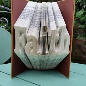 May include: A book folded into the shape of the word "Faith". The book is open and the pages are folded to create the letters. The book is sitting on a light blue table.