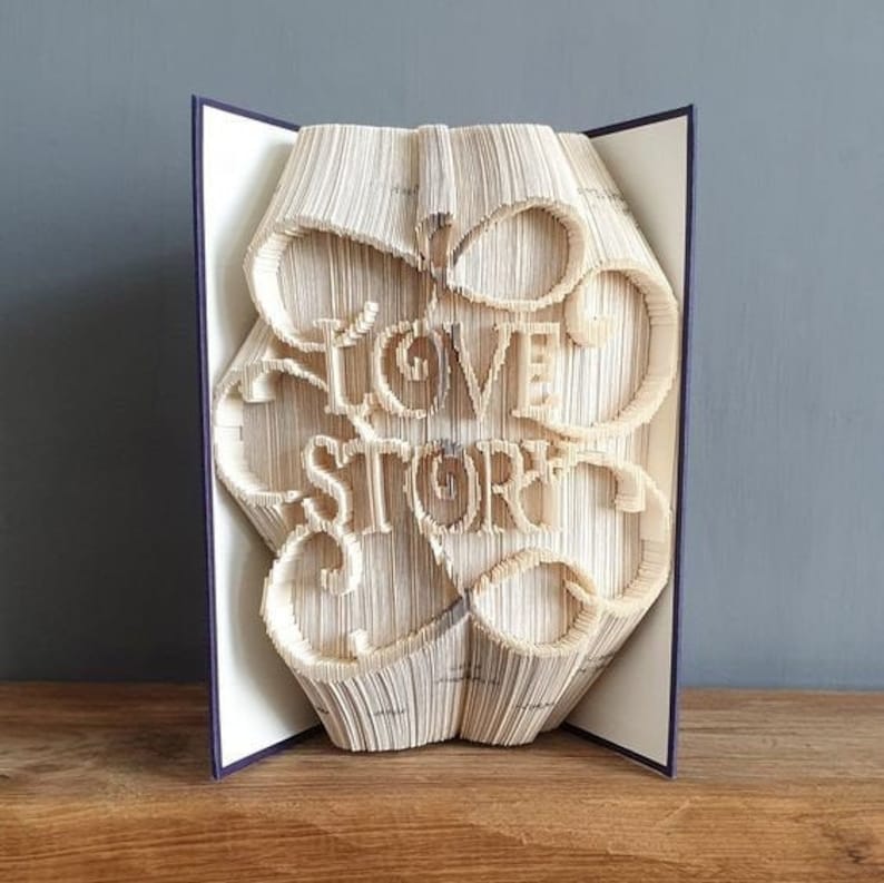Op de afbeelding: Een boek met de woorden "Love Story" gevouwen in de pagina's. Het boek is open en staat rechtop op een houten oppervlak.