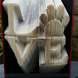 May include: A book art sculpture of the word "LOVE" folded from the pages of a book. The book is bound in a red cover with a black ribbon.