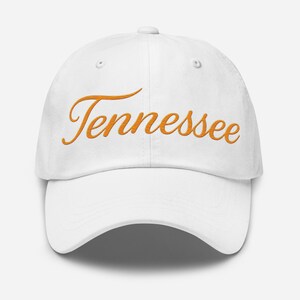 Puede incluir: Gorra de béisbol blanca con la palabra "Tennessee" bordada en escritura naranja. La gorra tiene una visera curva y un diseño de seis paneles.