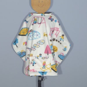 May include: White fleece poncho with a colorful print of children playing on a playground. The poncho has a pocket on the front.