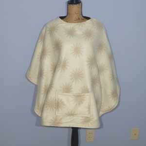 May include: A cream-colored fleece poncho with a brown starburst pattern. The poncho has a single pocket on the front.