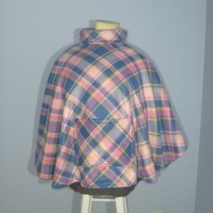 May include: A blue, pink, and yellow plaid fleece poncho with a pocket on the front. The poncho is on a white wooden stool.
