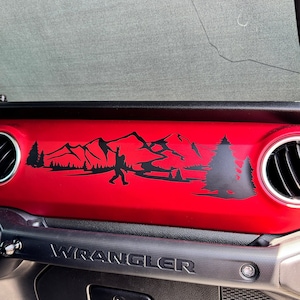 May include: A red Jeep Wrangler dashboard with a black mountain and forest scene decal. The decal features a silhouette of a person hiking in the mountains. The dashboard also has a black "WRANGLER" emblem.