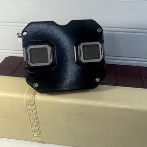 May include: A black View-Master stereoscope viewer resting on a rectangular, two-toned storage case. The case features a cream-colored front panel with the embossed text View-Master and a dark maroon lid and base, constructed from molded plastic.