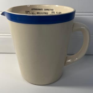 May include: A cream-colored ceramic measuring pitcher with a blue rim. The interior rim features black text reading APPROXIMATE CAPACITIES, CUPS, MILLILITRES, PTS, and FL OZS. It has a single handle on the right side and a small pouring spout.