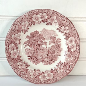 May include: A round, antique-style plate with a white center and a red floral and landscape design. The rim features a repeating floral pattern. The plate's center depicts a pastoral scene with trees, figures, and a building.