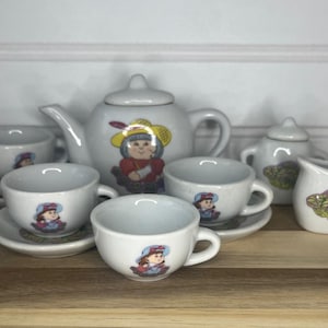 May include: A white ceramic tea set featuring a teapot, creamer, sugar bowl, and several teacups with saucers. Each piece is decorated with a cartoon character in a bonnet. The set is arranged on a wooden surface.