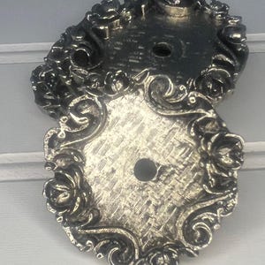May include: Two ornate, silver-colored cabinet hardware backplates. Each plate features a floral and scroll design around the edges, with a central hole for a screw. The plates have a textured surface.