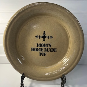 May include: A round, tan-colored ceramic pie dish with a glossy finish. The center of the dish features a black decorative heart-shaped icon and the black printed text "MOM'S HOME MADE PIE" in a bold, sans-serif font. The dish has a wide, smooth rim.