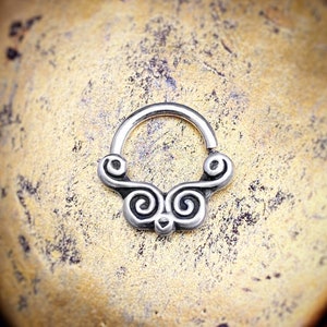 May include: A silver-colored septum ring with an ornate, symmetrical design. The decorative element features swirling patterns and a central detail. The jewelry is shown against a textured, golden-brown background.