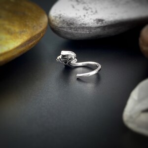 May include: A silver nose ring with a small, detailed design on the front. The ring is curved and has a small opening for insertion.