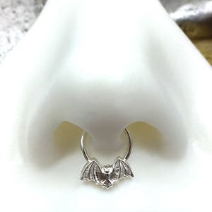 May include: Silver nose ring with a bat charm. The ring is a hoop style and the bat is detailed with wings and a body.