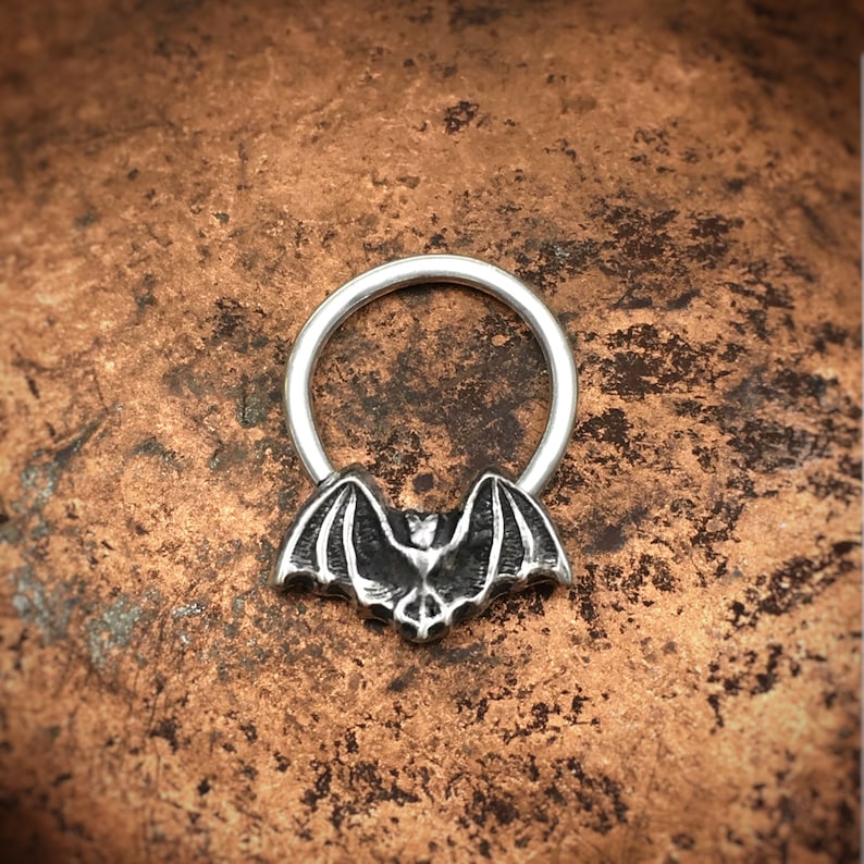 May include: Silver septum ring with a bat design. The bat is detailed and has a textured finish.