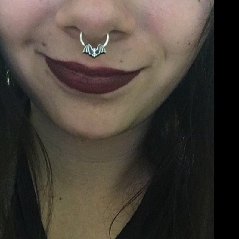 May include: A close-up of a person's nose with a silver septum ring featuring a bat design.