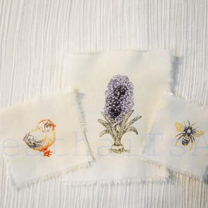 May include: Three small, off-white fabric squares with embroidered designs. One features a chick, another a purple hyacinth, and the third a bee. The edges are frayed, and the background is a textured white fabric.