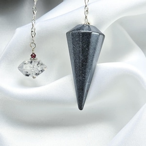 May include: Two pendulums hang from silver chains against a white background. One is a faceted, dark gray stone. The other is a clear, diamond-shaped crystal with inclusions and a small red bead. The pendulums are likely used for divination.
