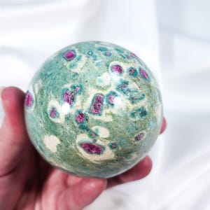Peut inclure: Une sphère de pierre précieuse avec un motif moucheté de turquoise, crème et magenta. La pierre polie est tenue dans une main, mettant en valeur sa surface lisse et les couleurs vives du minéral. La sphère est un objet décoratif.