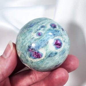 Peut inclure: Une sphère de pierre précieuse polie avec un motif moucheté de turquoise, de bleu et de blanc. La sphère est parsemée d'inclusions magenta profondes. La pierre est tenue dans une main, mettant en valeur sa forme lisse et arrondie.