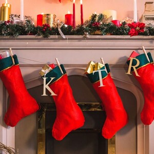 May include: Four red Christmas stockings with green and gold trim are hung on a white fireplace mantel. Each stocking has a wooden letter attached to it, spelling out the word "J K T R".