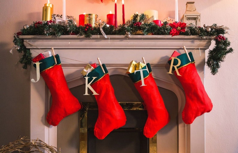 May include: Four red Christmas stockings with green and gold trim are hung on a white fireplace mantel. Each stocking has a wooden letter attached to it, spelling out the word "J K T R".