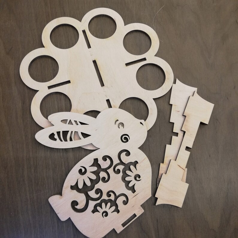 May include: A wooden cutout of a rabbit holding a lantern with a floral design. The rabbit is surrounded by a circle of eight smaller circles, also cut out of wood.