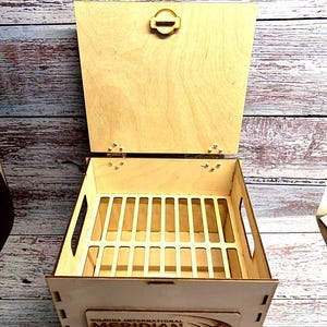 May include: A light-coloured wooden box with a hinged lid and a rectangular shape. The box has a slotted interior and a decorative label with text. The lid is secured with a wooden clasp.
