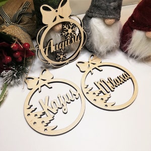 May include: Three wooden Christmas ornaments with names "Augusta", "Kaijus", and "Milana" written on them. Each ornament features a bow and a design of a Christmas tree with stars.