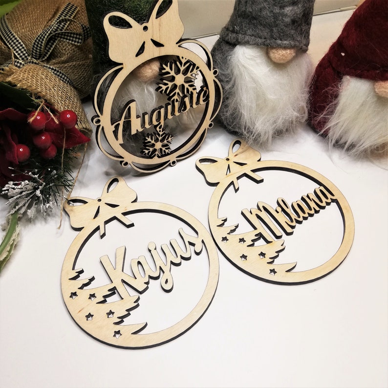 May include: Three wooden Christmas ornaments with names "Augusta", "Kaijus", and "Milana" written on them. Each ornament features a bow and a design of a Christmas tree with stars.