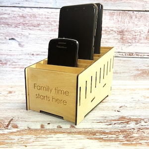 Könnte beinhalten: Holztelefonständer mit Schlitzen für mehrere Telefone. Der Ständer hat den Text "Family time starts here" auf der Vorderseite eingraviert.