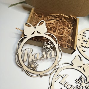 May include: Three unfinished wooden Christmas ornaments with a bow and snowflake designs. The ornaments are personalized with the names "Rasa", "Anya", and "Lily".