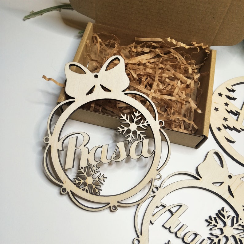 May include: Three unfinished wooden Christmas ornaments with a bow and snowflake designs. The ornaments are personalized with the names "Rasa", "Anya", and "Lily".