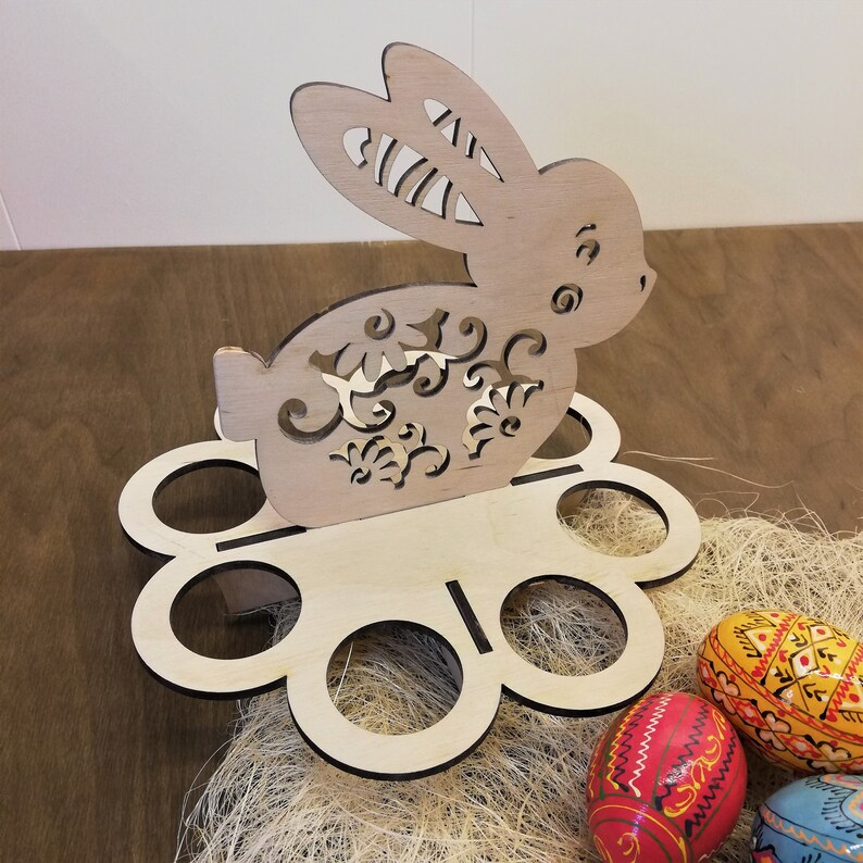 May include: Wooden Easter egg holder shaped like a bunny with intricate cutouts. The bunny is holding a flower design. The holder has six egg slots and is sitting on a bed of shredded paper. There are three painted Easter eggs in the foreground.