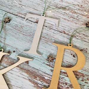 May include: Three wooden letters, "K", "R", and "T", are hanging from twine with small wooden snowflakes. The "K" is natural wood, the "R" is gold, and the "T" is silver.