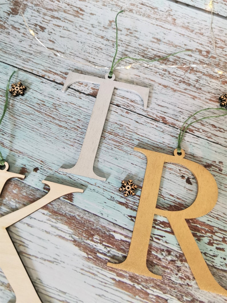 May include: Three wooden letters, "K", "R", and "T", are hanging from twine with small wooden snowflakes. The "K" is natural wood, the "R" is gold, and the "T" is silver.