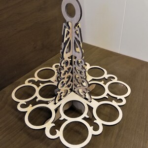 May include: A wooden egg holder with a decorative design. It has a handle at the top and eight slots for eggs. The holder is made of light brown wood with a darker brown stain.